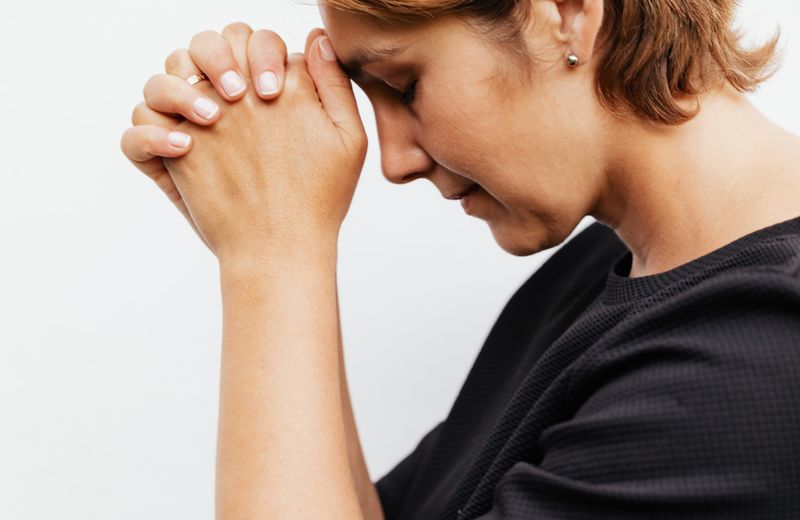 Mulher de olhos fechados e mãos unidas, em um momento de fé.